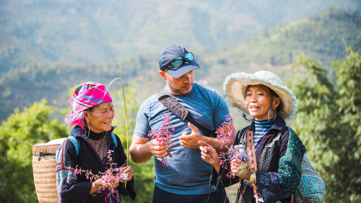 white european male tourist standing together with two indigenous woman from the black hmong tribe in the sapa mountains in vietnam, holding wild flowers in hands and talking