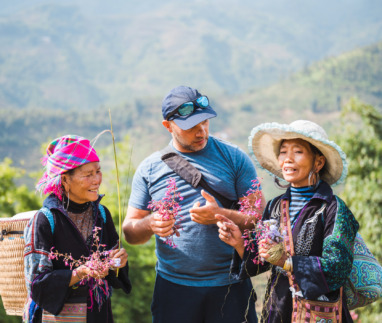 white european male tourist standing together with two indigenous woman from the black hmong tribe in the sapa mountains in vietnam, holding wild flowers in hands and talking