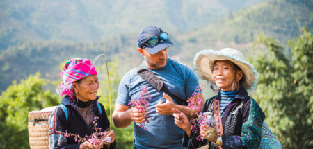 white european male tourist standing together with two indigenous woman from the black hmong tribe in the sapa mountains in vietnam, holding wild flowers in hands and talking