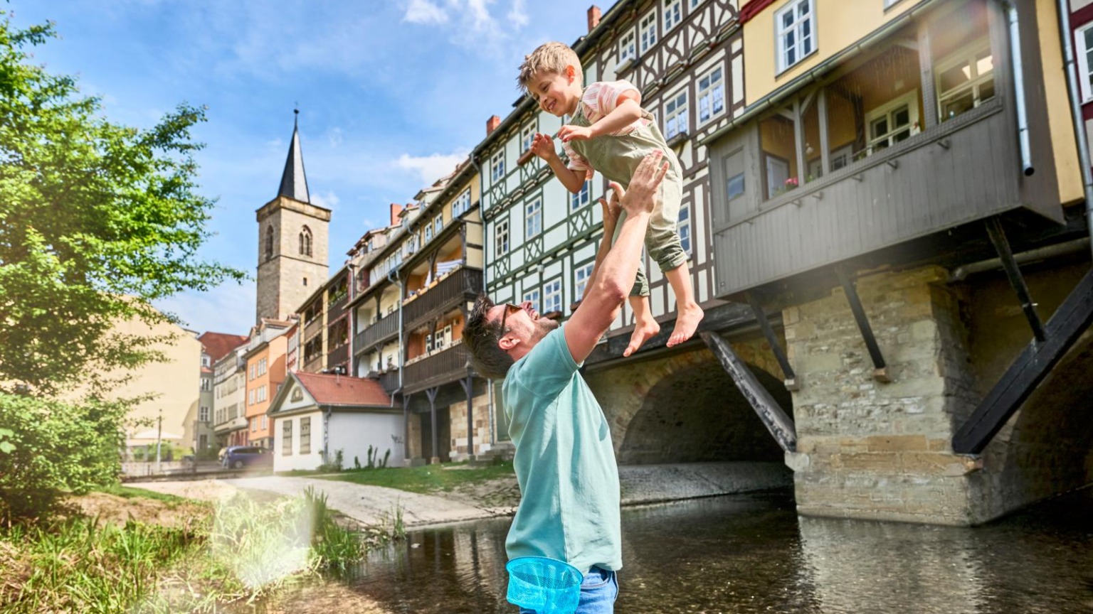Vader tilt kind op bij de Krämerbrücke in Erfurt, met vakwerkhuizen boven de rivier de Gera.