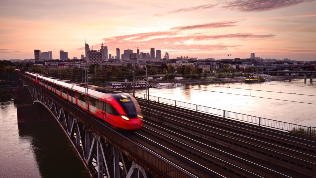 Sneltrein rijdt over brug bij zonsondergang in Warschau met skyline op de achtergrond.