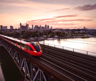 Sneltrein rijdt over brug bij zonsondergang in Warschau met skyline op de achtergrond.