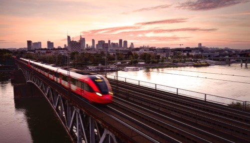 Sneltrein rijdt over brug bij zonsondergang in Warschau met skyline op de achtergrond.