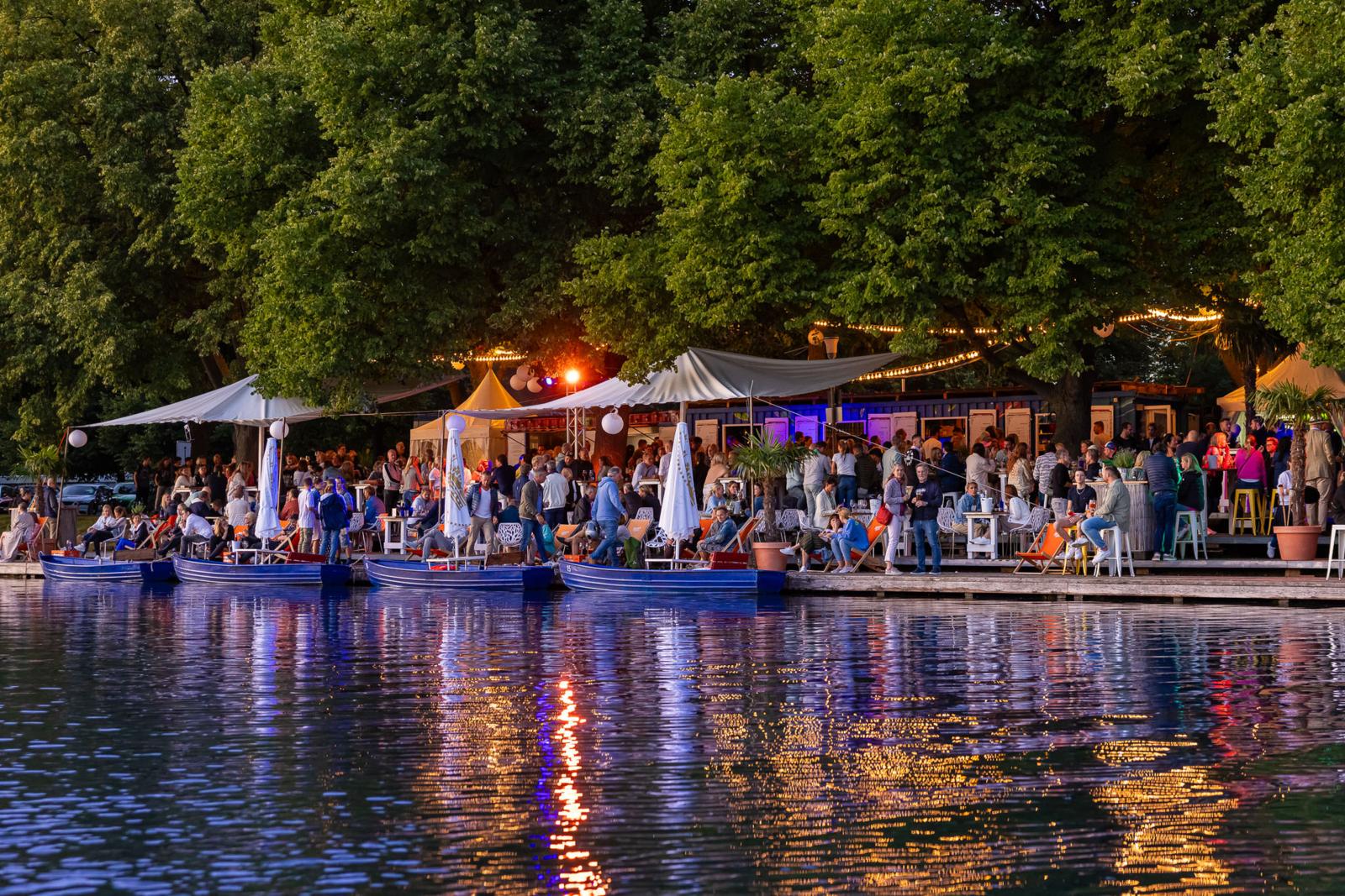 Drukke zomeravond aan het water in Hannover, Duitsland, met mensen op terrasboten en sfeervolle verlichting.