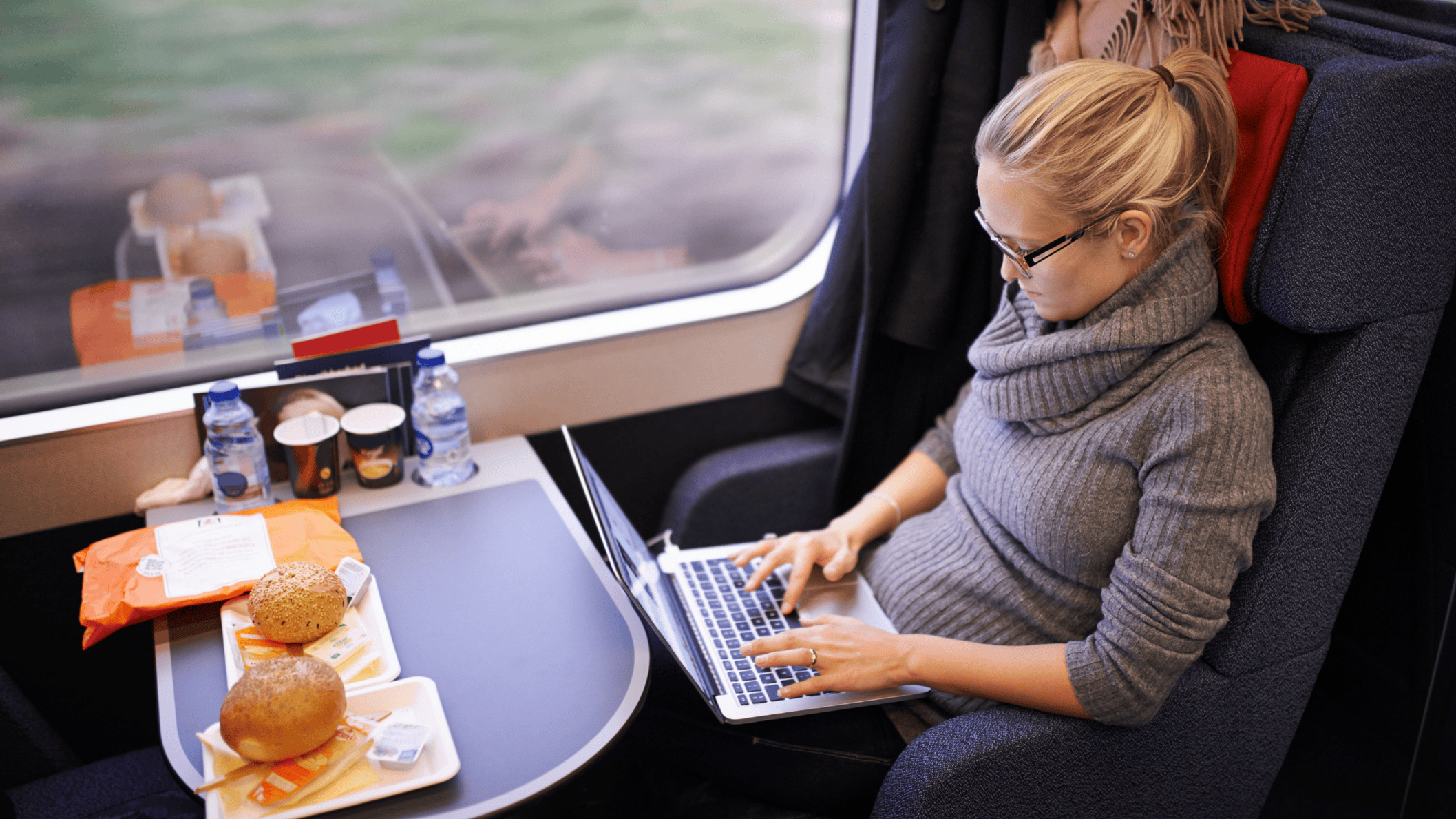 Vrouw werkt op laptop terwijl ze met de trein reist, met ontbijt en koffie op tafel.