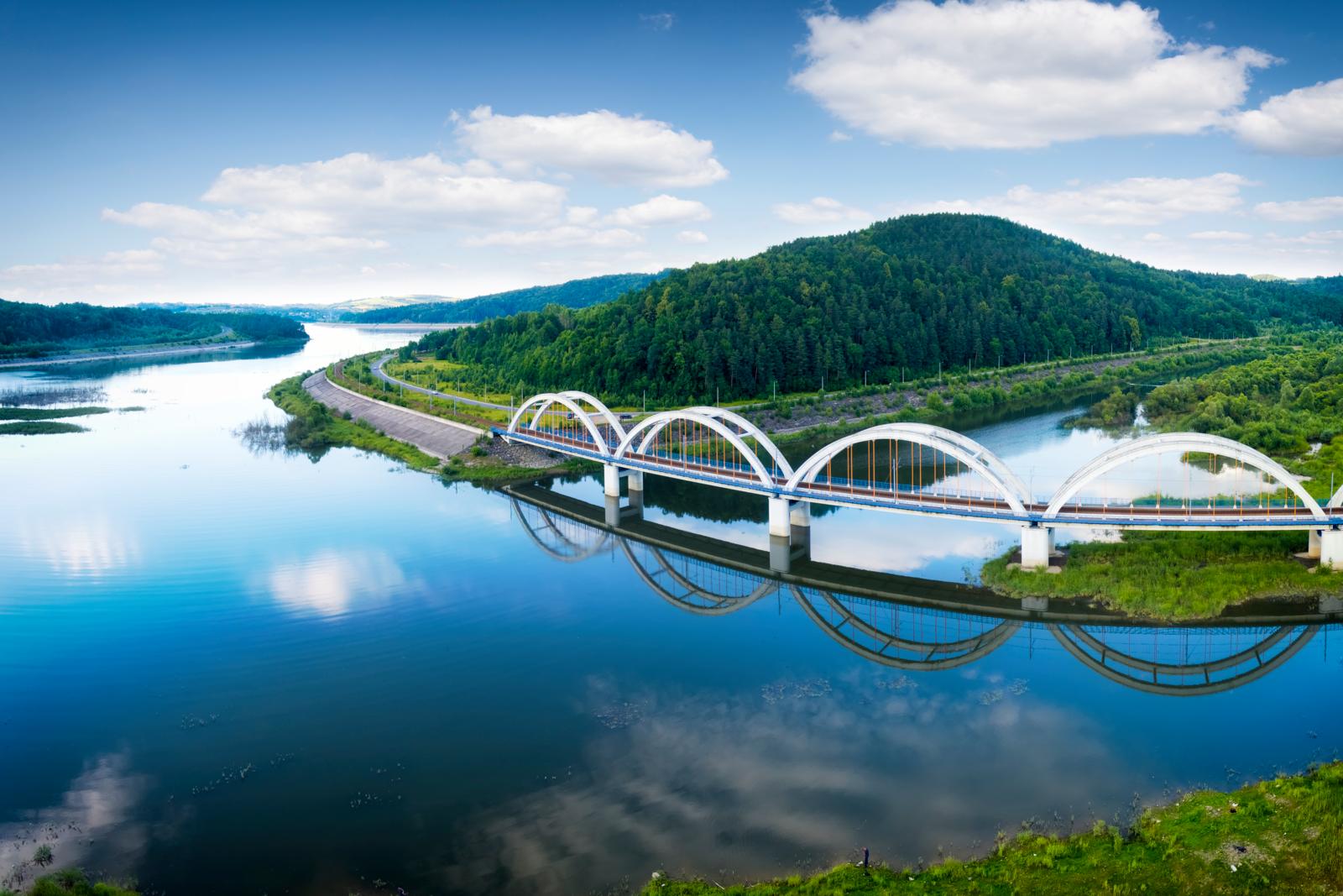 Trein rijdt over een witte boogbrug bij Sucha Beskidzka in Polen, boven de spiegelende rivier de Skawa en omringd door groene heuvels.