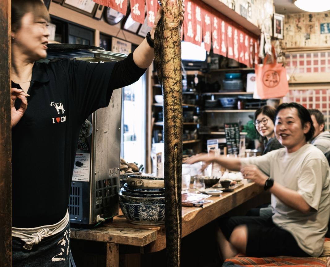 Gastvrouw toont een grote vis in Kamontei, een traditioneel restaurant in Kōchi, Shikoku, Japan, met gasten aan de bar