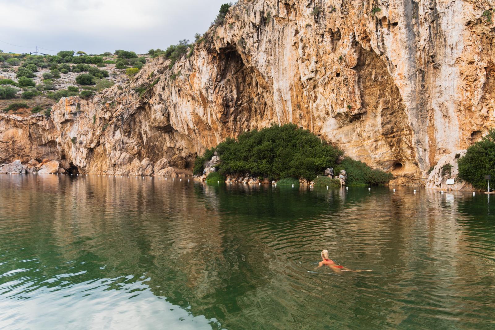 Vrouw zwemt in het groene thermale water van Lake Vouliagmeni bij Athene, omringd door rotsen.