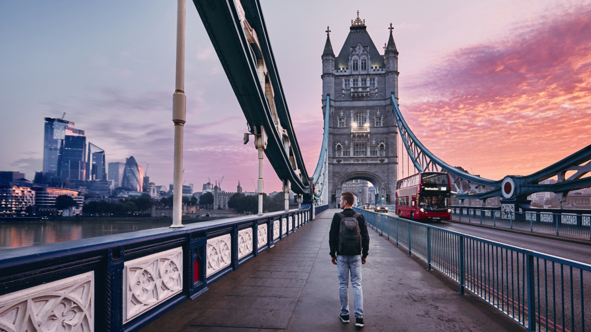 Jongeman met rugzak loopt over de Tower Bridge in Londen, Engeland tijdens zonsopkomst met wolkenkrabbers op de achtergrond