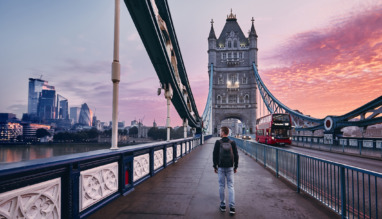 Jongeman met rugzak loopt over de Tower Bridge in Londen, Engeland tijdens zonsopkomst met wolkenkrabbers op de achtergrond