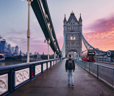 Jongeman met rugzak loopt over de Tower Bridge in Londen, Engeland tijdens zonsopkomst met wolkenkrabbers op de achtergrond