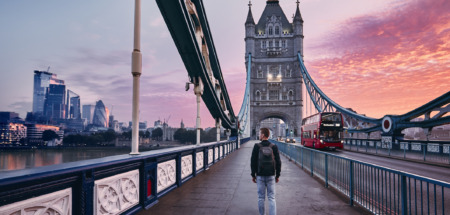 Jongeman met rugzak loopt over de Tower Bridge in Londen, Engeland tijdens zonsopkomst met wolkenkrabbers op de achtergrond