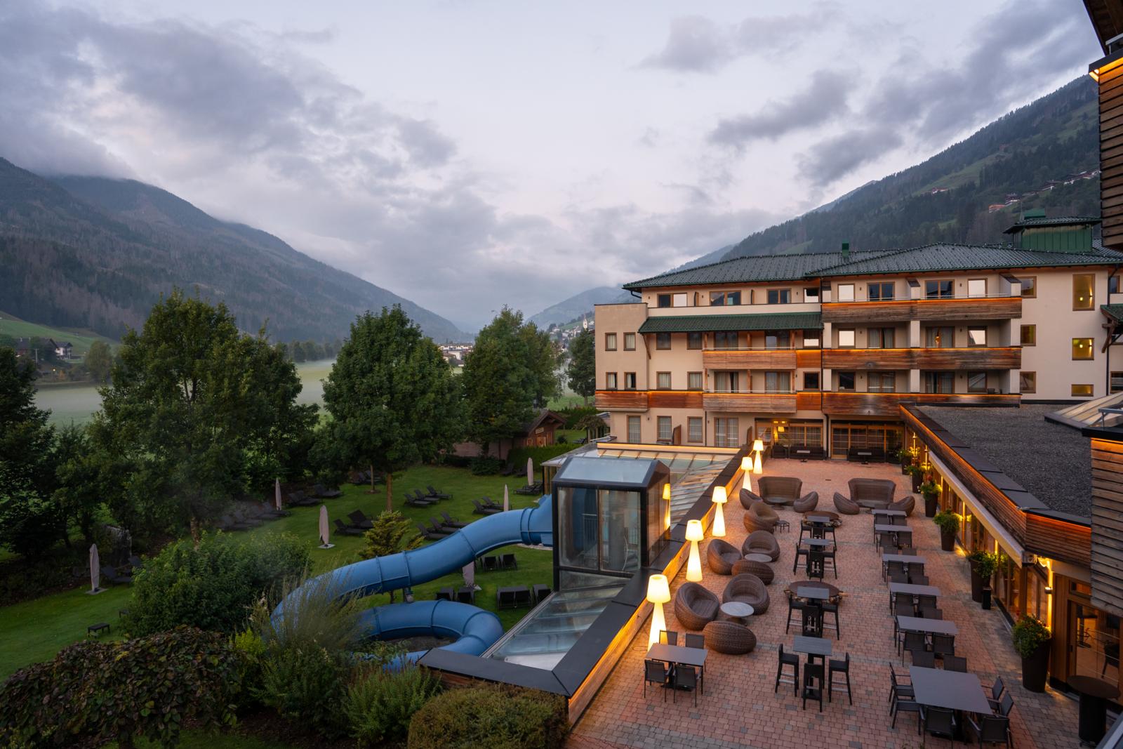 Hotel met terras, glijbaan en tuin in een bergvallei in Oost-Tirol bij ochtendlicht.
