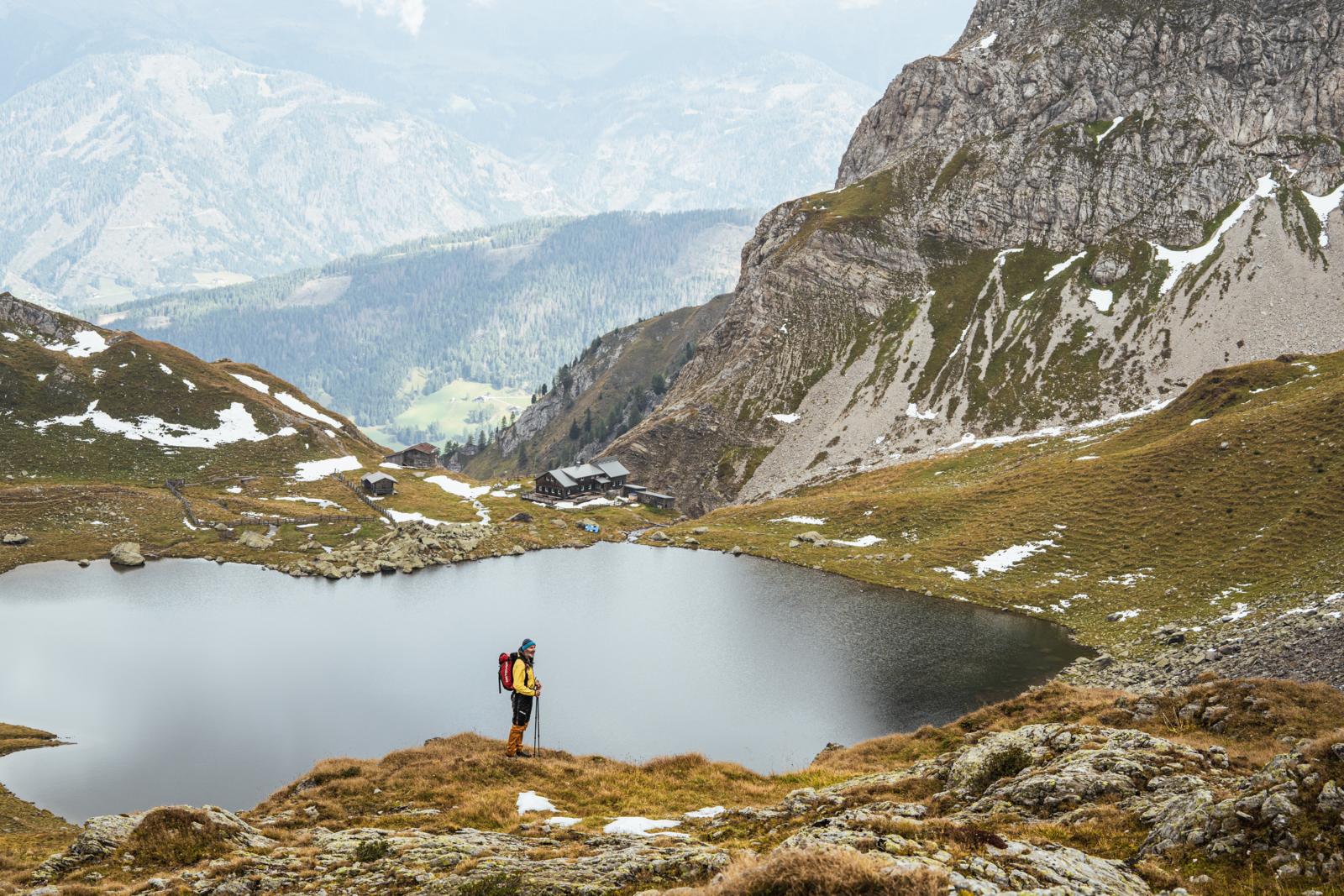 Wandelaar bij een bergmeer in Oost-Tirol met uitzicht op ruige pieken en almhutten in de verte.