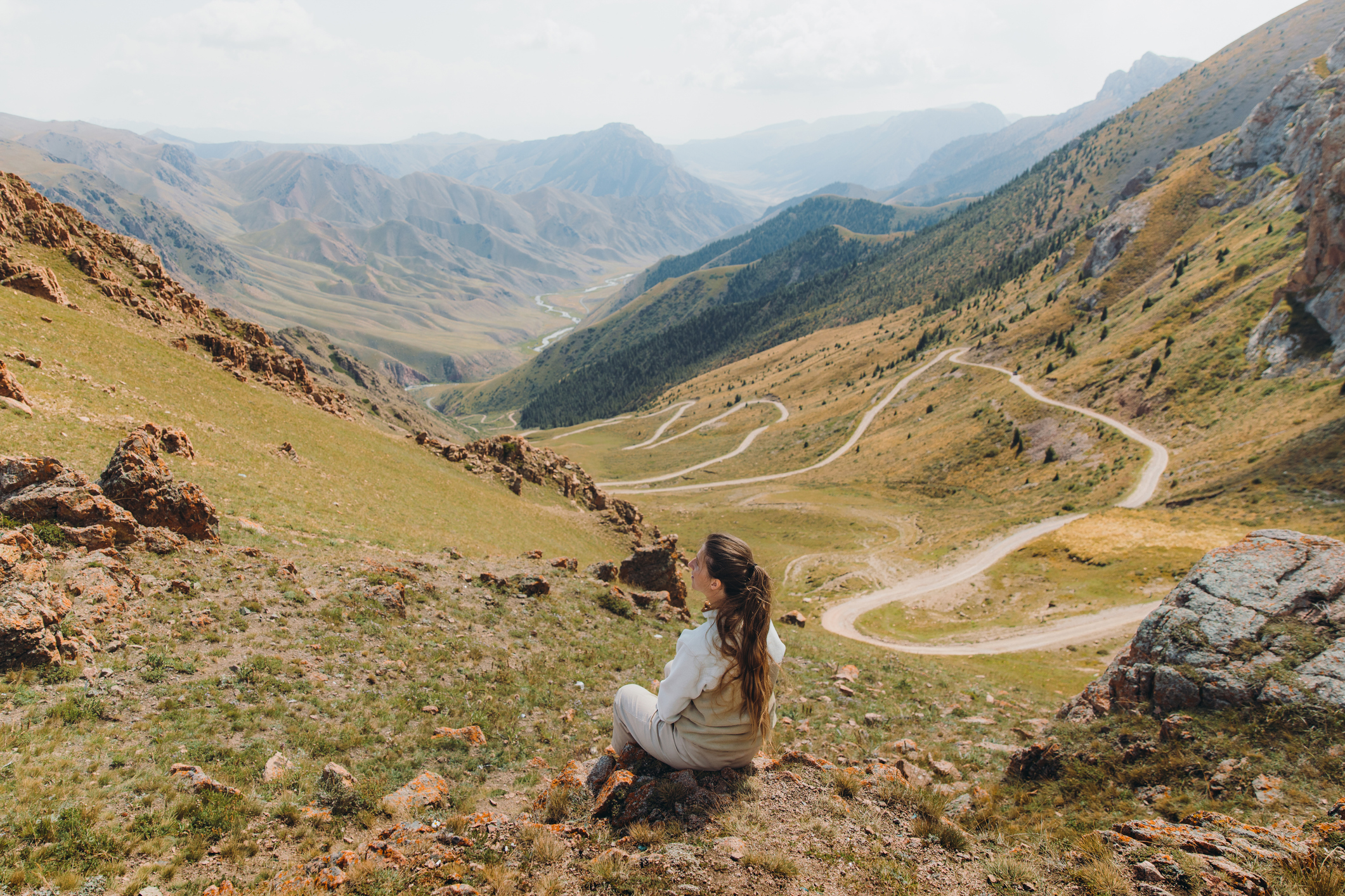 Vrouw zit op een berghelling en kijkt uit over een slingerende bergweg en groene valleien in Tadzjikistan.