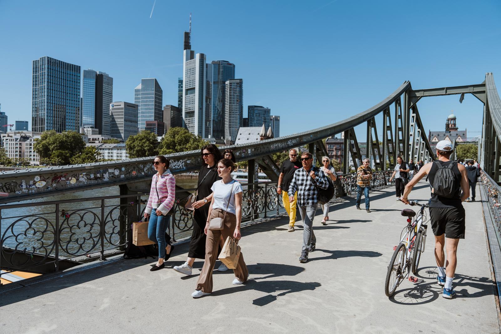 Wandelaars en fietsers op de Eiserner Steg met de skyline van Frankfurt op de achtergrond.