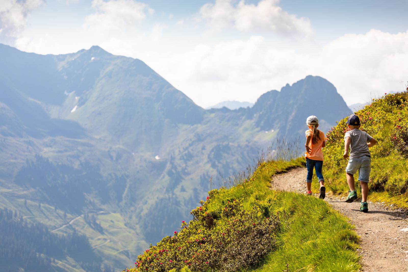 Twee kinderen wandelen over een bergpad met uitzicht op groene Alpenvalleien.