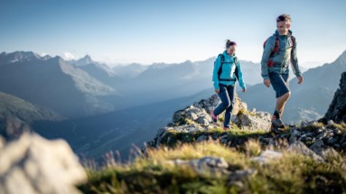 Twee wandelaars lopen over een bergkam met uitzicht op de Alpen.