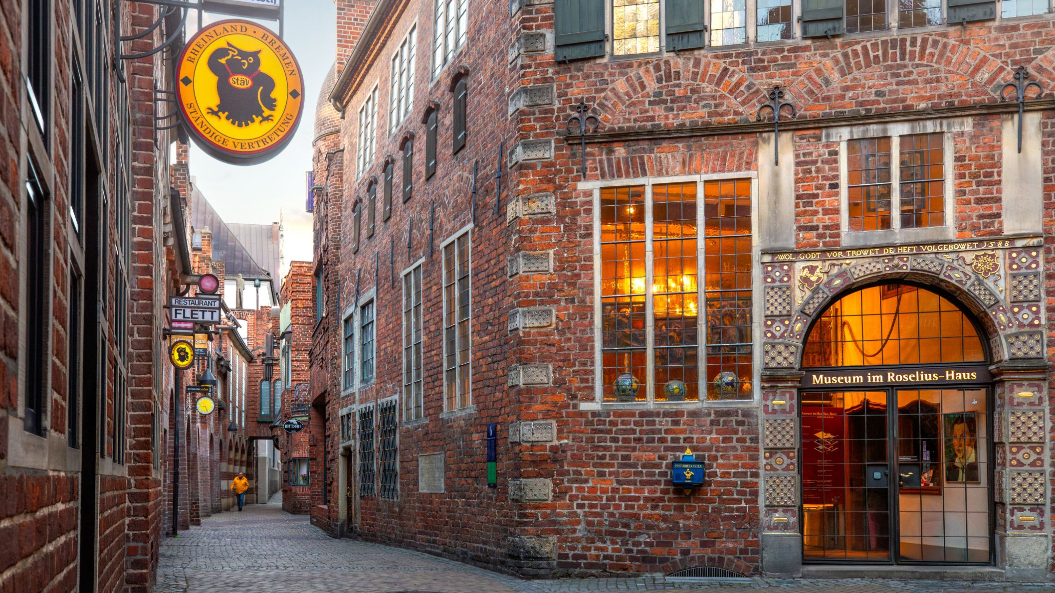Sfeervolle opname van de smalle Böttcherstraße in Bremen met de bakstenen gevel van het Roselius-Haus en karakteristieke uithangborden bij warm avondlicht.