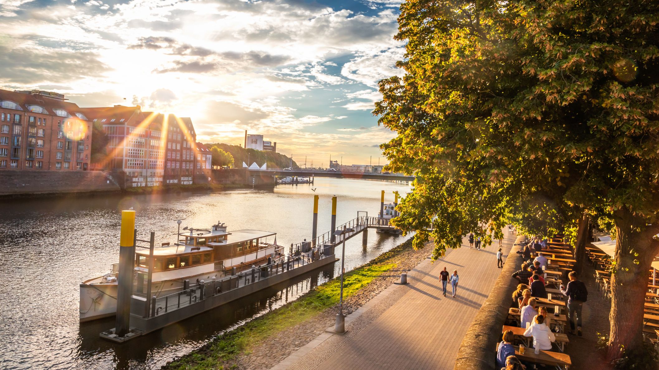 Panoramisch uitzicht op de Schlachte-promenade in Bremen bij zonsondergang met terrasjes onder de bomen en een aangemeerde boot op de Wezer.