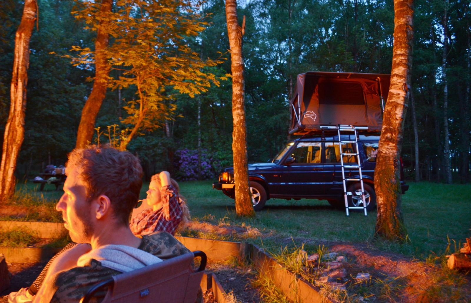 Auto met daktent op camping in bosrijke omgeving bij kampvuur.
