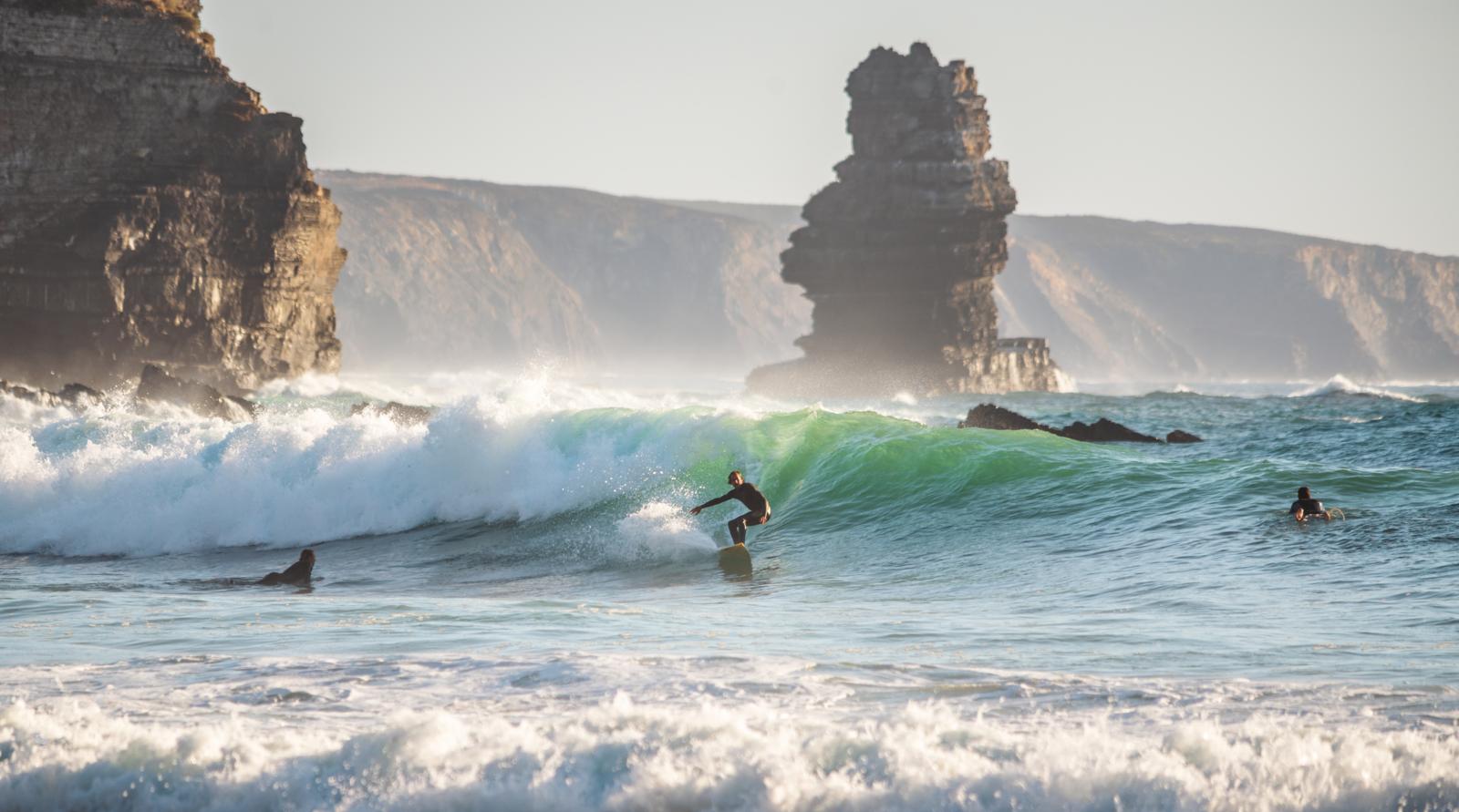 Twee surfers met surfplanken staan op rotsen langs een ruige Atlantische kust.