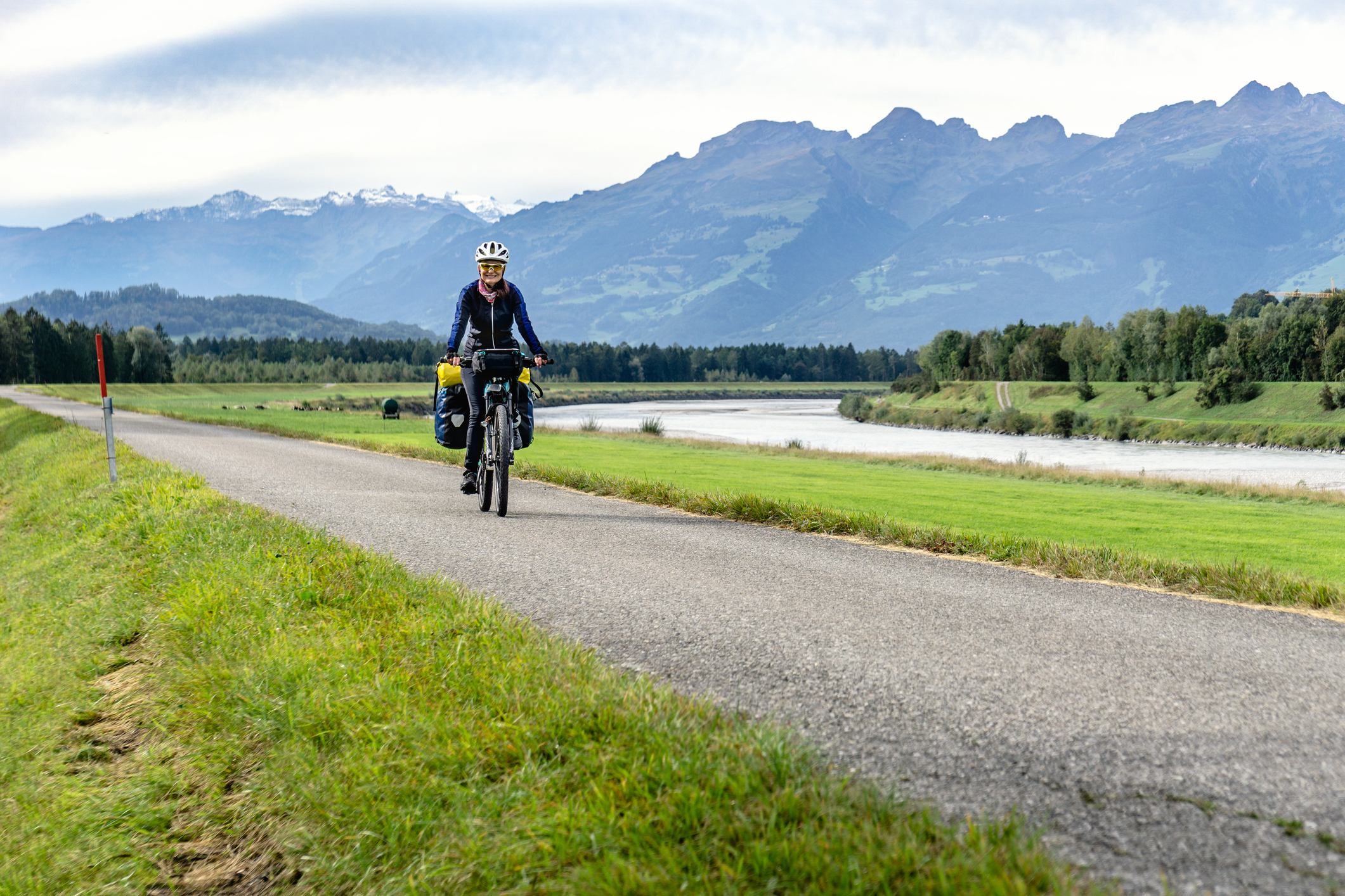 Fietser met bepakking op een fietspad langs een rivier met de Alpen op de achtergrond.