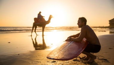 Surfer met surfboard en kameel op het strand van Cable Beach bij zonsondergang in Broome, Australië.