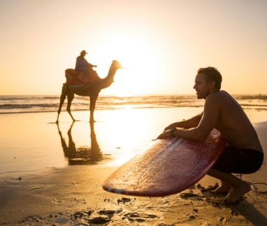 Surfer met surfboard en kameel op het strand van Cable Beach bij zonsondergang in Broome, Australië.