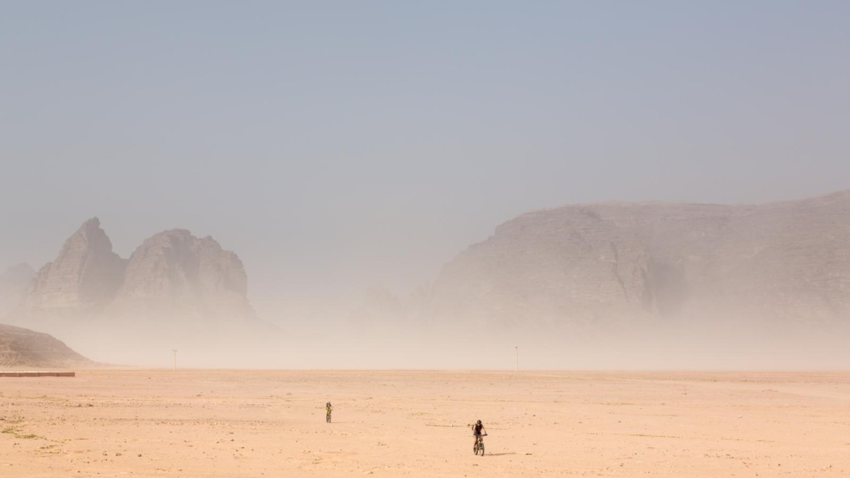 Twee fietsers in de woestijn van Wadi Rum met zandstorm en rotsformaties op de achtergrond.