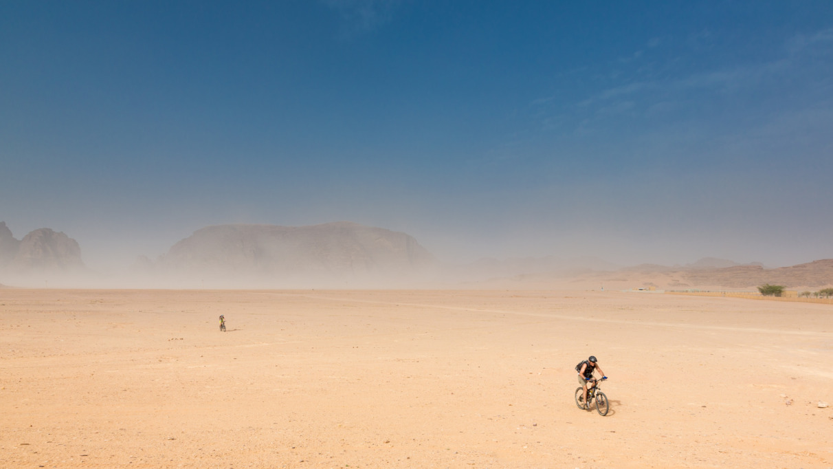 Twee fietsers in de woestijn van Wadi Rum in Jordanie met zandstorm en rotsformaties op de achtergrond.