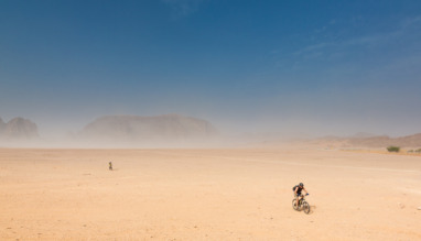 Twee fietsers in de woestijn van Wadi Rum in Jordanie met zandstorm en rotsformaties op de achtergrond.