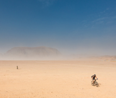 Twee fietsers in de woestijn van Wadi Rum in Jordanie met zandstorm en rotsformaties op de achtergrond.
