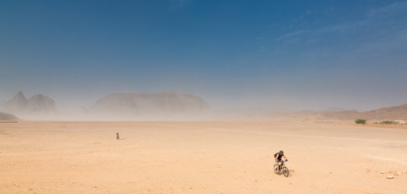 Twee fietsers in de woestijn van Wadi Rum in Jordanie met zandstorm en rotsformaties op de achtergrond.