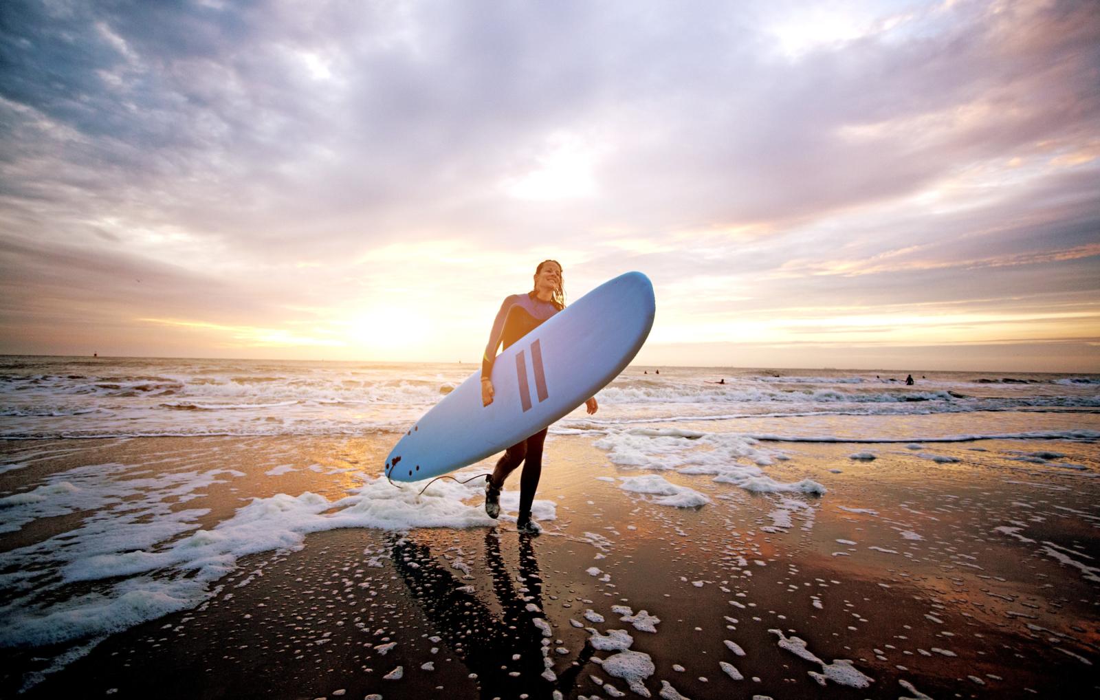Surfer met surfboard loopt uit zee bij zonsondergang Nederlandse kust.