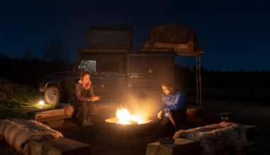 Stel zit bij kampvuur op de Veluwe met safarijeep met daktent op achtergrond