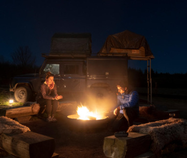 Stel zit bij kampvuur op de Veluwe met safarijeep met daktent op achtergrond