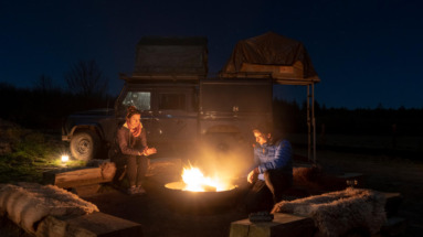 Stel zit bij kampvuur op de Veluwe met safarijeep met daktent op achtergrond
