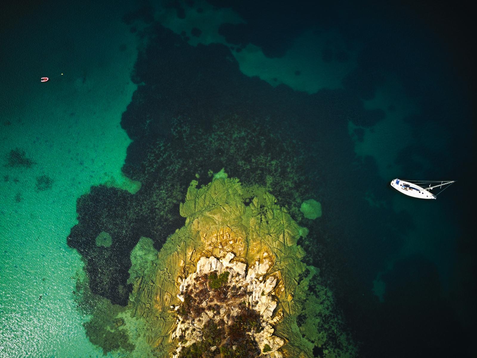 Dronefoto van een klein rotsachtig eiland met zeilboot in helder turquoise water.