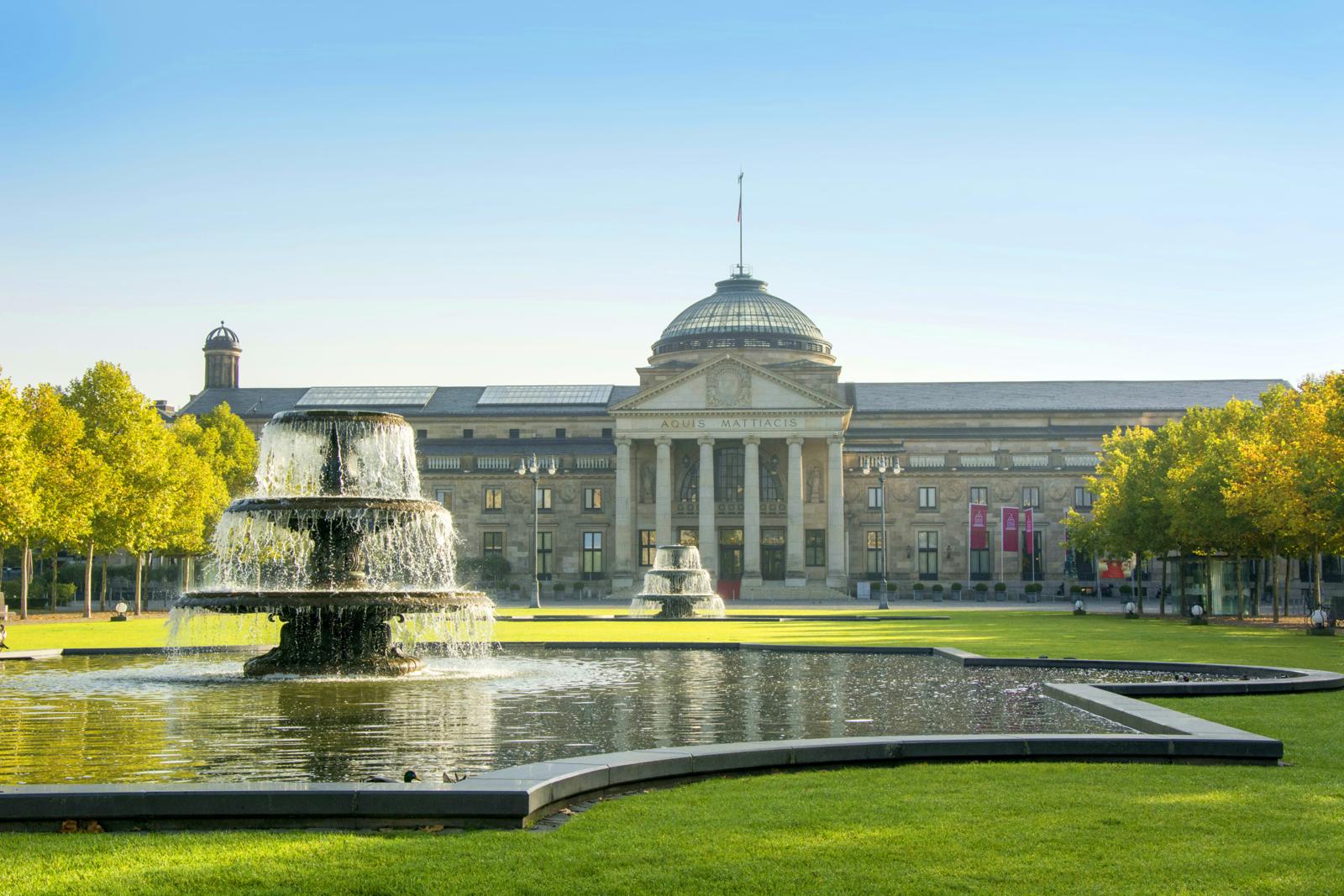 Vooraanzicht van het neoclassicistische Kurhaus in Wiesbaden met een grote fontein en groen gazon.