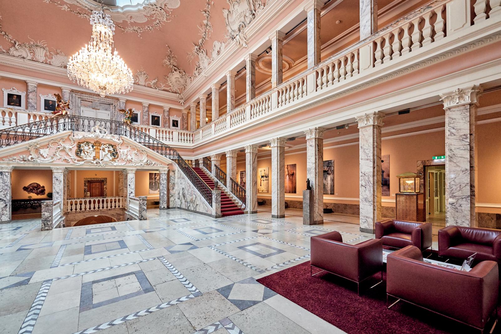 Een luxe klassieke hal met marmeren zuilen, een kristallen kroonluchter en een imposante trap met rode loper.
