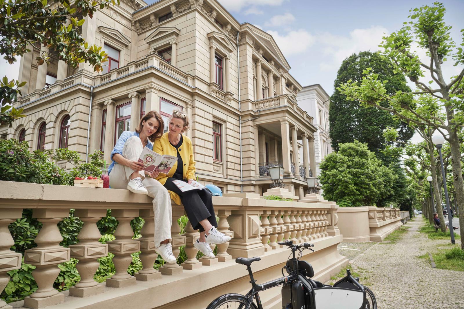 Twee vrouwen bekijken een stadskaart zittend op een stenen balustrade voor een imposante klassieke villa in Wiesbaden.