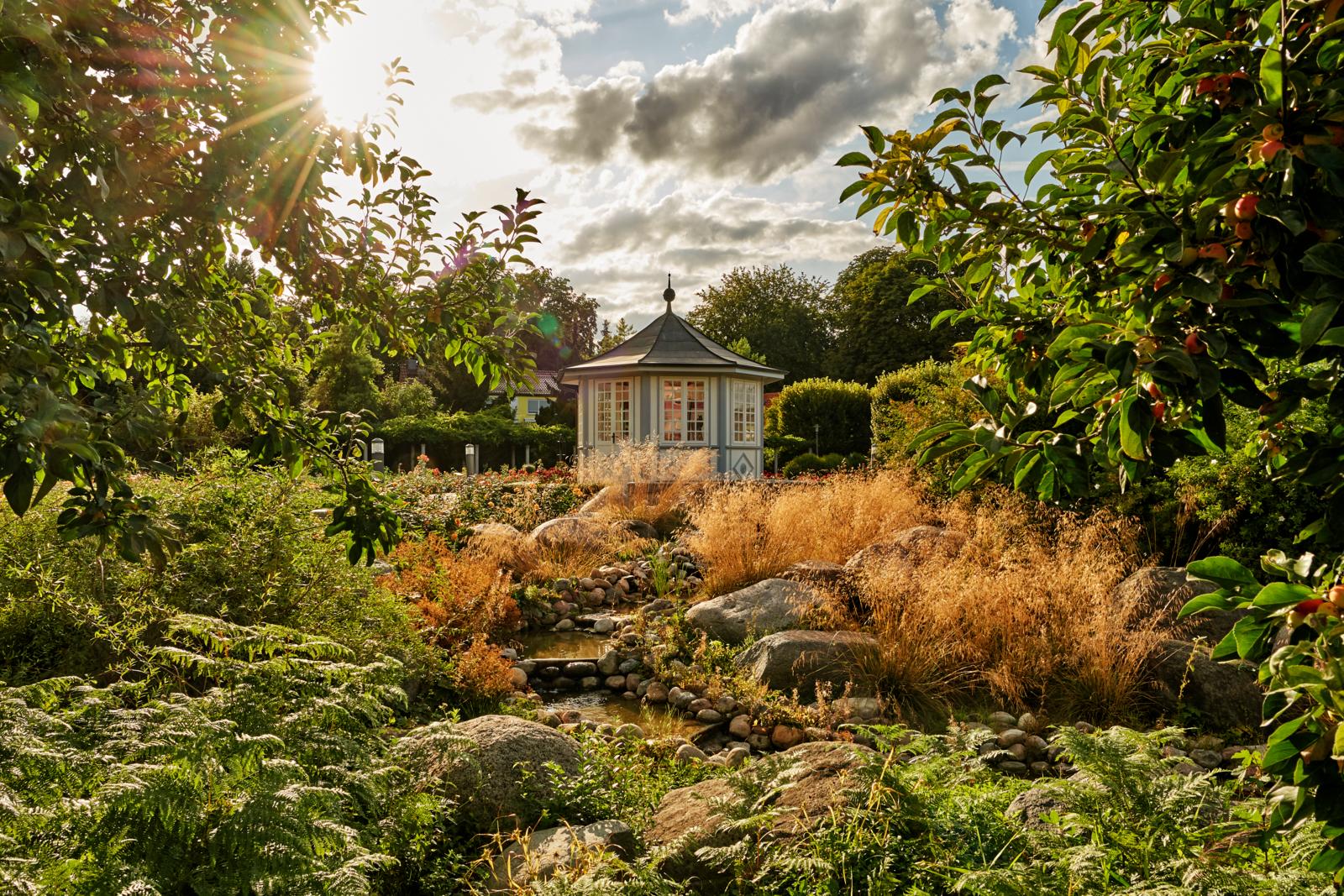 Een romantisch paviljoen in een weelderige rozentuin met rotsen en een beekje bij tegenlicht.