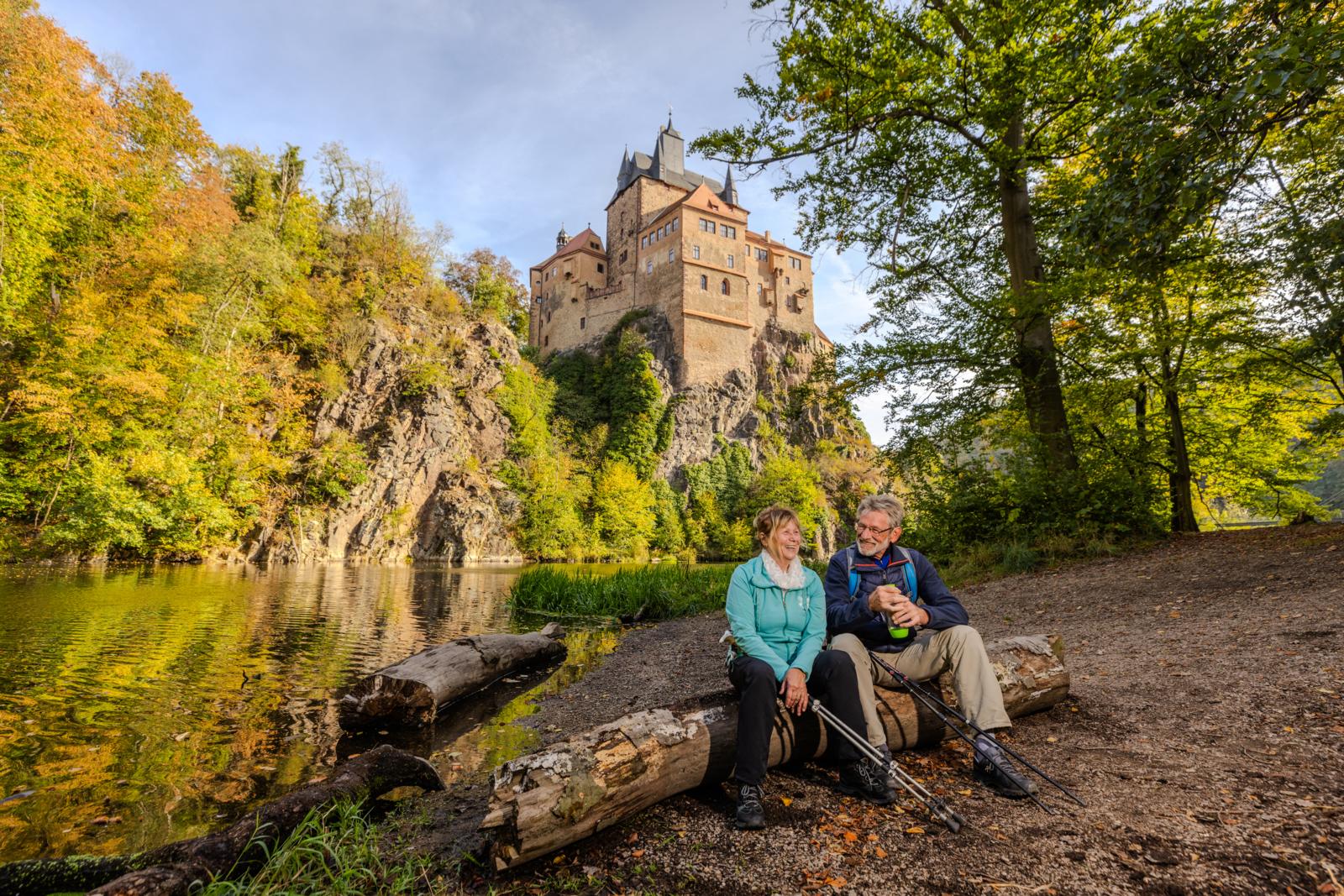 Twee wandelaars rusten uit op een boomstam bij het water met uitzicht op de hooggelegen Burg Kriebstein.