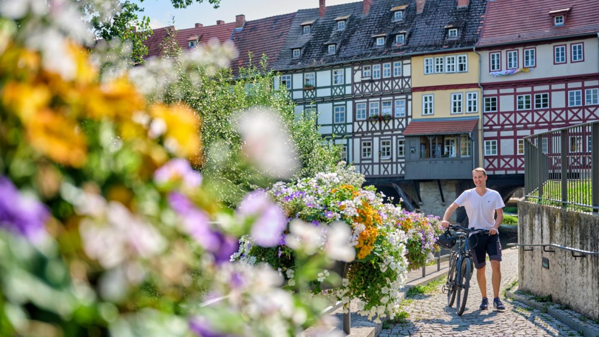 Een fietser wandelt over een kasseienpad langs kleurrijke bloemen met op de achtergrond de historische vakwerkhuizen van de Krämerbrücke.