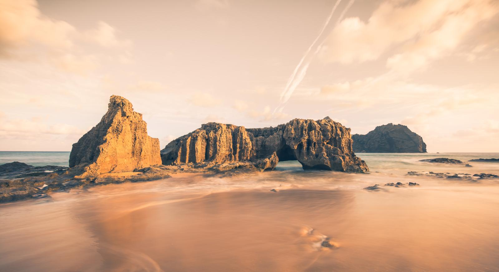 Een lange-sluitertijd landschapsfoto toont de iconische natuurlijke rotsboog en rotsnaalden op een zandstrand in Asturië bij golden hour, met een zijdeachtige zee en dramatische wolkenlucht.