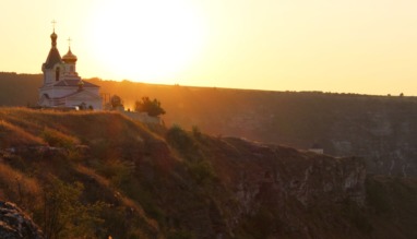 Een wit-orthodoxe kerk met gouden koepels staat op de rand van een steile kalkstenen klif tijdens een warme, gouden zonsondergang in Orheiul Vechi.