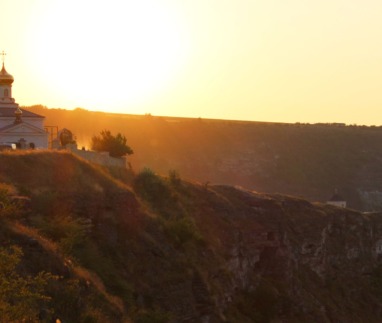 Een wit-orthodoxe kerk met gouden koepels staat op de rand van een steile kalkstenen klif tijdens een warme, gouden zonsondergang in Orheiul Vechi.