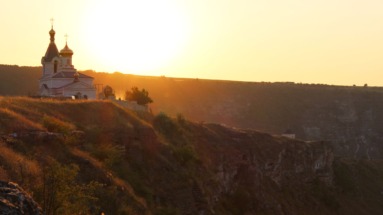 Een wit-orthodoxe kerk met gouden koepels staat op de rand van een steile kalkstenen klif tijdens een warme, gouden zonsondergang in Orheiul Vechi.