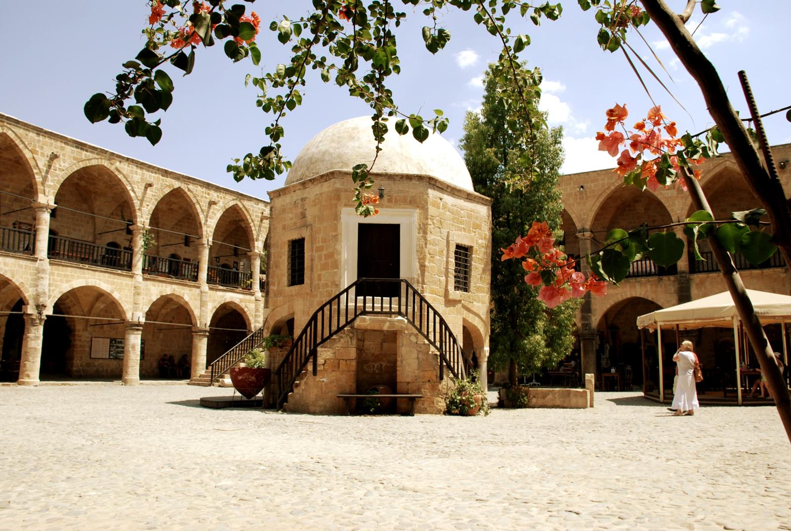 De binnenplaats van de Buyuk Han in Nicosia met zijn karakteristieke bogen, centrale moskee met koepel en bloeiende bougainvillea onder een blauwe lucht.